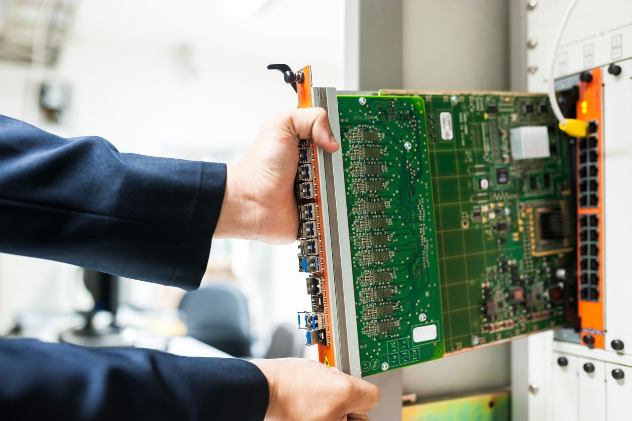 Two hands in dark blue sleeves sliding a green circuit board with a row of network ports into an orange and white equipment chassis