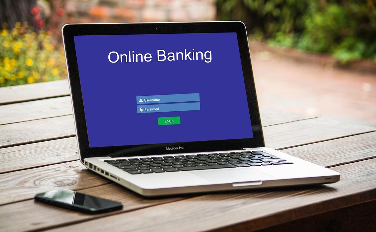 A laptop showing an online banking login screen with username and password fields, a phone beside it on a wooden table - phishing pages imitate real logins like this to capture your credentials.